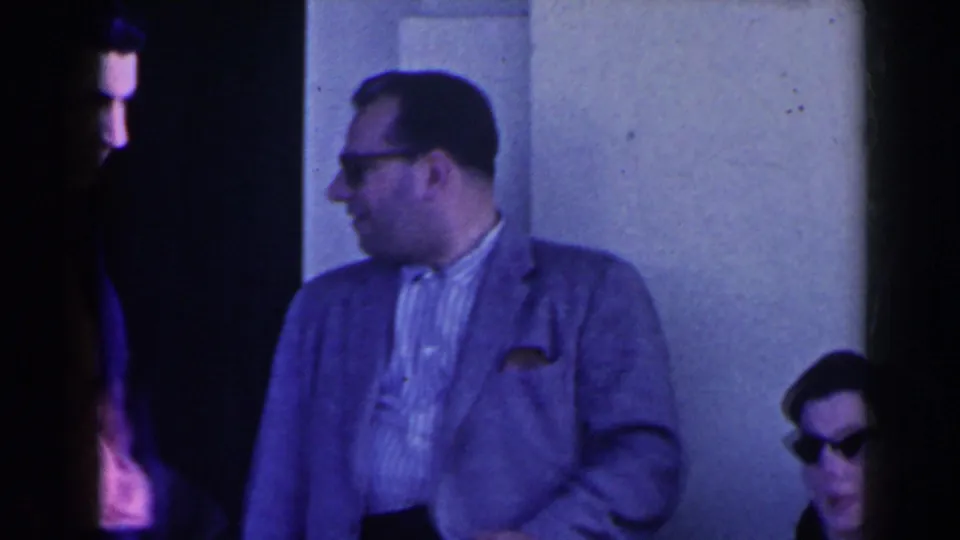1963 Niagara Falls home-movie frame with a man in a light jacket turning toward a seated woman near a building entrance.