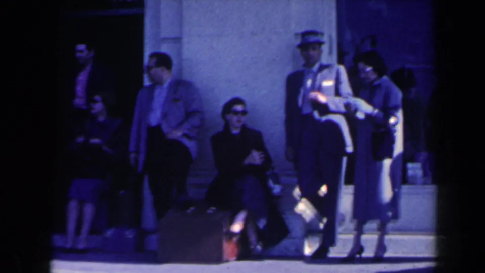 People in 1960s clothing wait outside a Niagara Falls building near steps, luggage, and a uniformed attendant.
