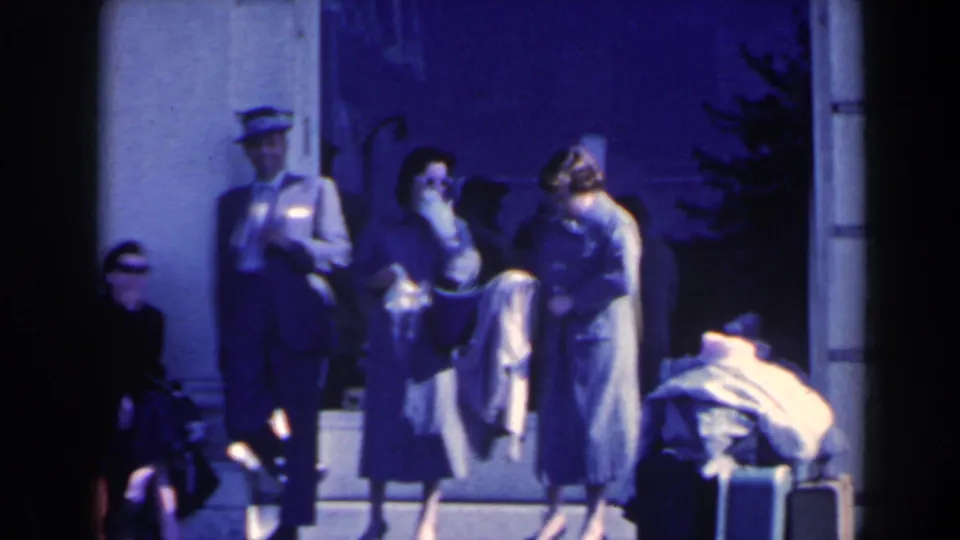 People in 1960s attire stand and sit outside a Niagara Falls building, with luggage and a uniformed attendant visible.