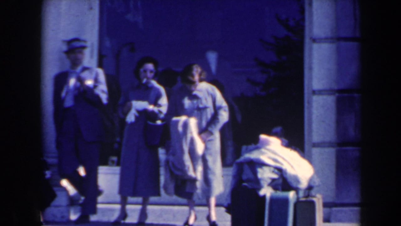 1963 8mm frame of adults at an entryway in Niagara Falls, Canada, with two women, suited men, and luggage in shade.