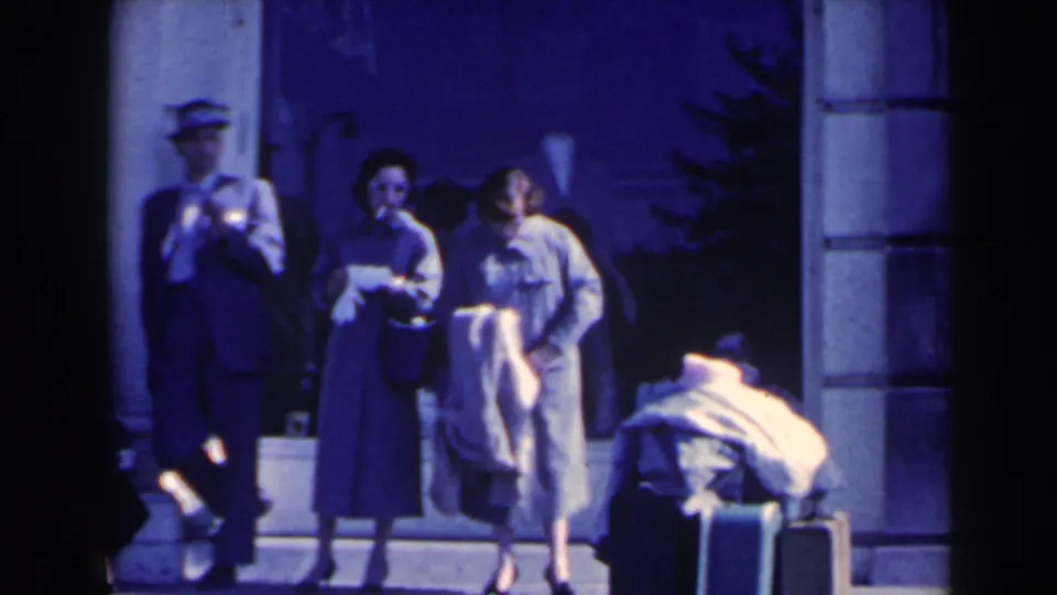 1963 8mm frame of adults at an entryway in Niagara Falls, Canada, with two women, suited men, and luggage in shade.
