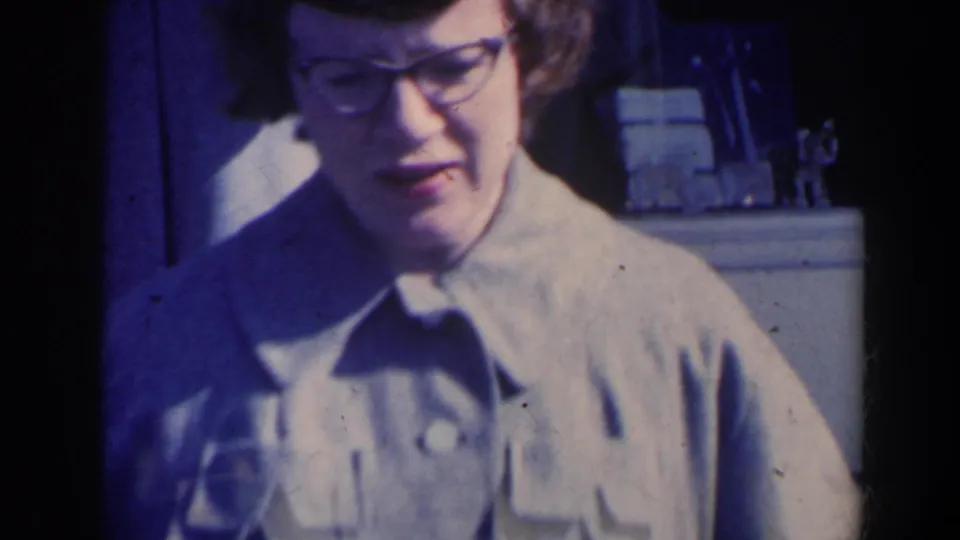 Close-up of a woman in glasses and a light jacket, with the entryway and other adults blurred behind her.