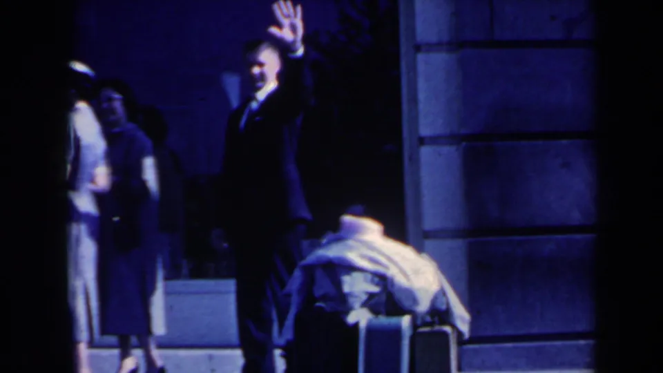 A suited man waves toward the camera near the shaded entrance, with luggage and other adults behind him.