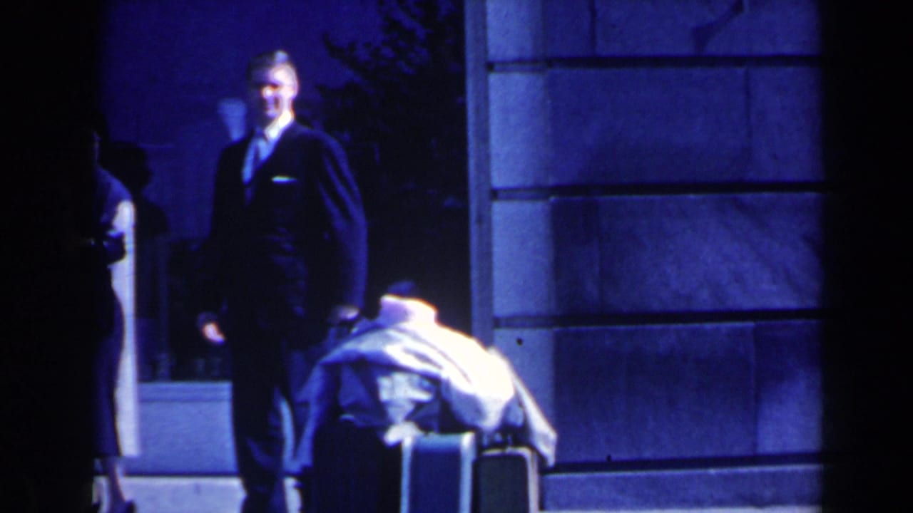 Man in a dark suit stands beside luggage near a stone building at night.