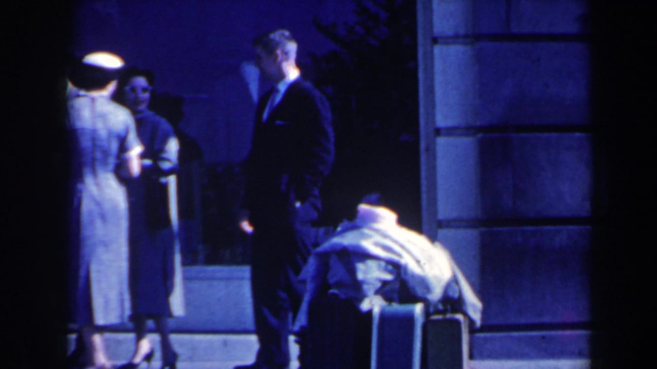 Two women and a suited man stand near luggage outside a building at night.