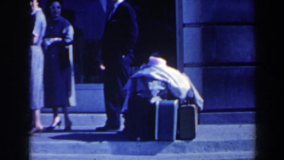 A woman in mid-century dress stands near a suited man and stacked luggage in a night exterior home-movie frame.