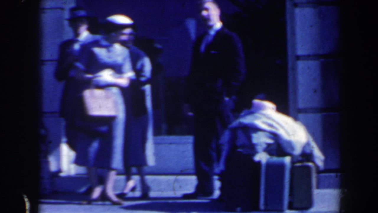 1963 Niagara Falls, Canada home-movie frame showing travelers in dresses, hats, and a suit waiting beside luggage near a station entrance.