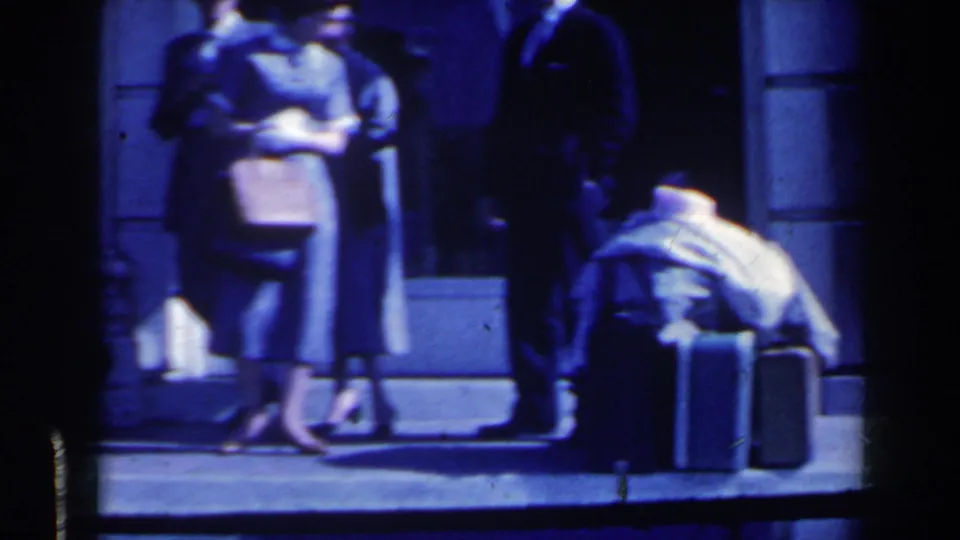 Travelers at a Niagara Falls, Canada bus station appear beside suitcases, with a woman in a dress and hat and a man in a suit visible.