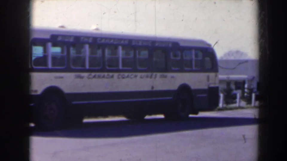 The Canada Coach Lines bus remains parked, with a low building and small distant figures visible beyond the road.