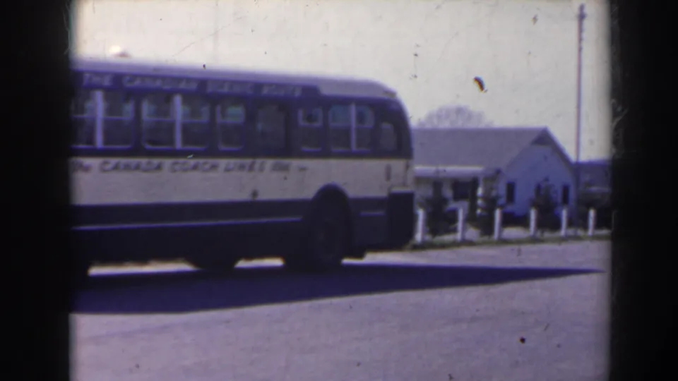 The rear section of the Canada Coach Lines bus passes across the frame, with a roadside building and small people in the background.
