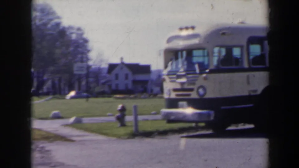 The bus is framed at the right edge beside a grassy roadside scene with a sidewalk, fire hydrant, trees, and a white house-like building.