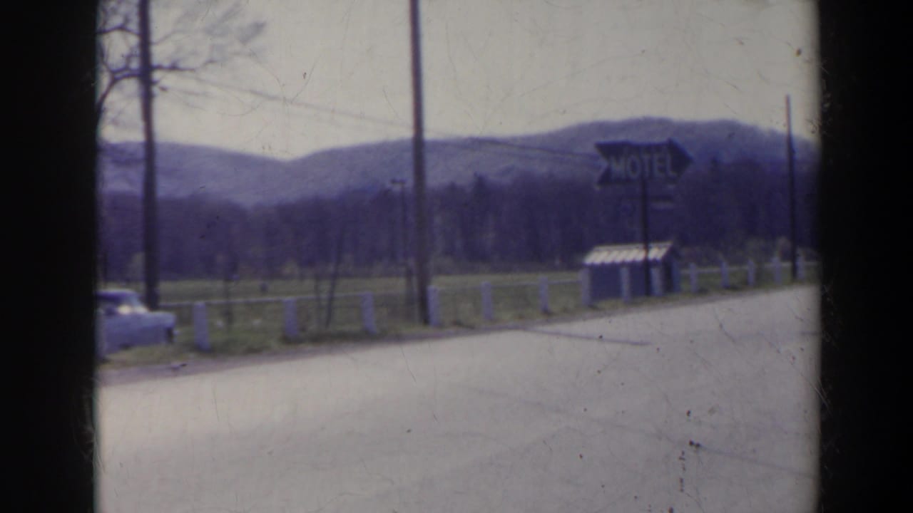 Roadside view with motel sign, road, utility poles, fencing, hills, and a passing car at frame edge.