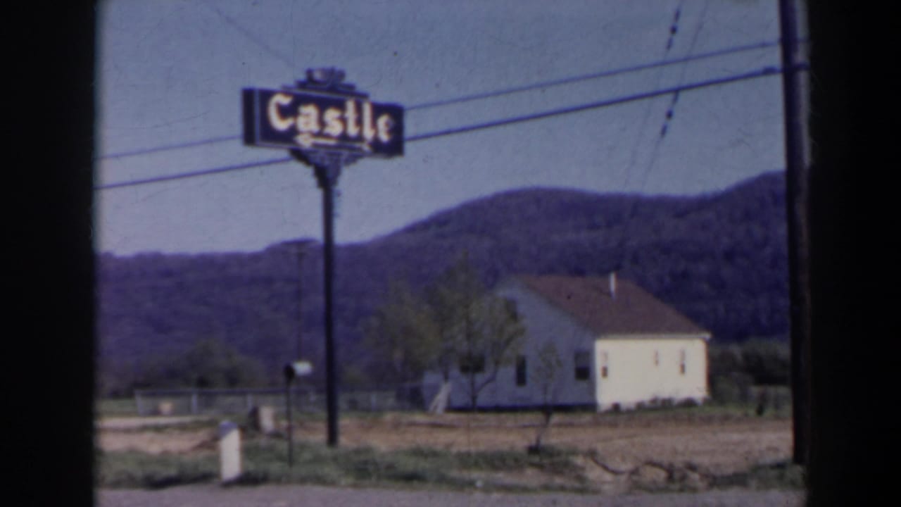 Castle sign on a pole beside a white building, mailbox, utility wires, and wooded hills.