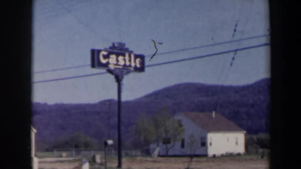 Closer 1963 8mm view of the Castle sign near Niagara Falls, Canada, framed against wires, wooded hills, a white building, and a mailbox.