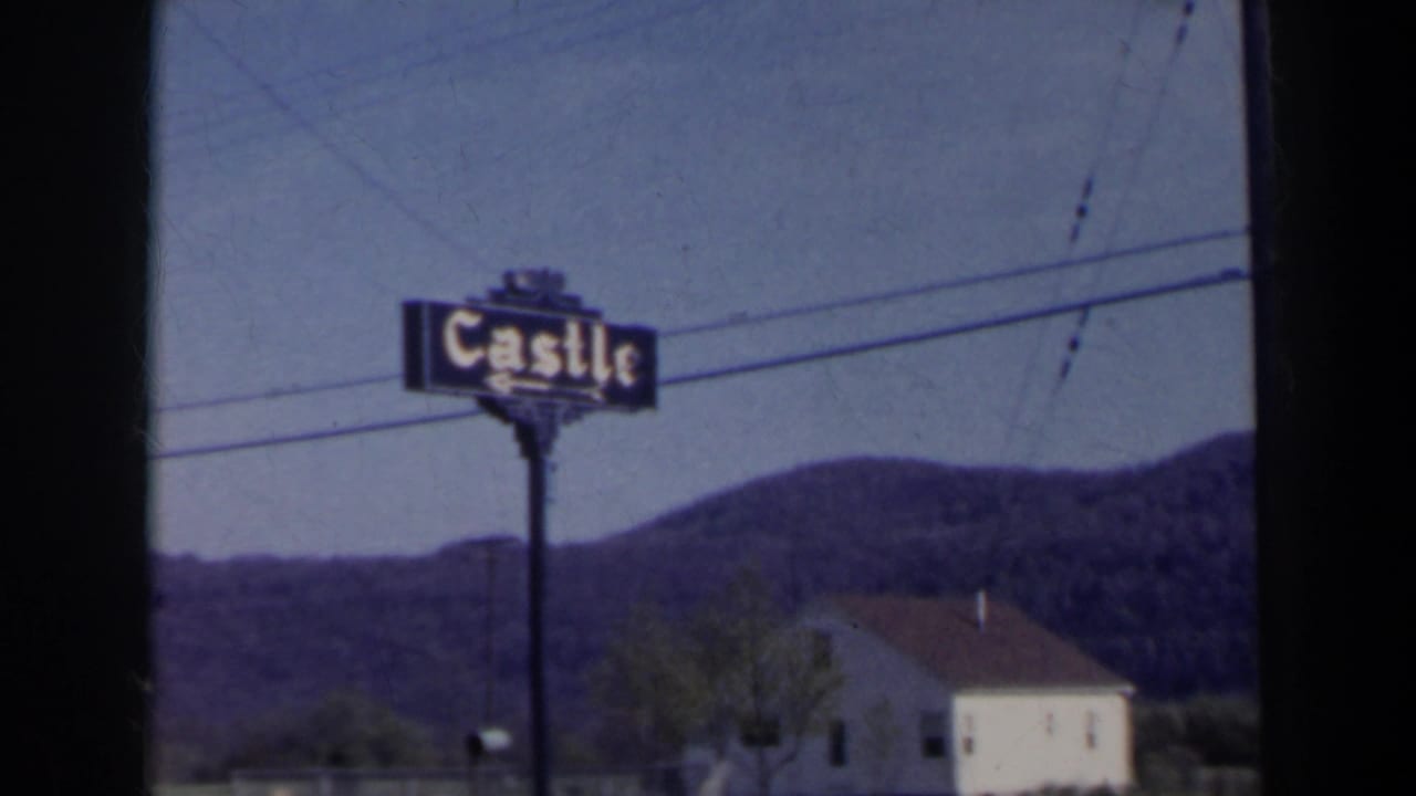 Castle roadside sign with hills, utility wires, and a pale building in the background.