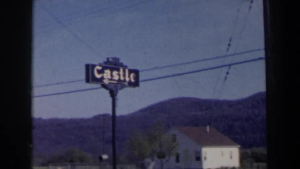 1963 Niagara Falls home-movie frame showing a Castle sign beside a road, with hills, overhead wires, and a light-colored building behind it.