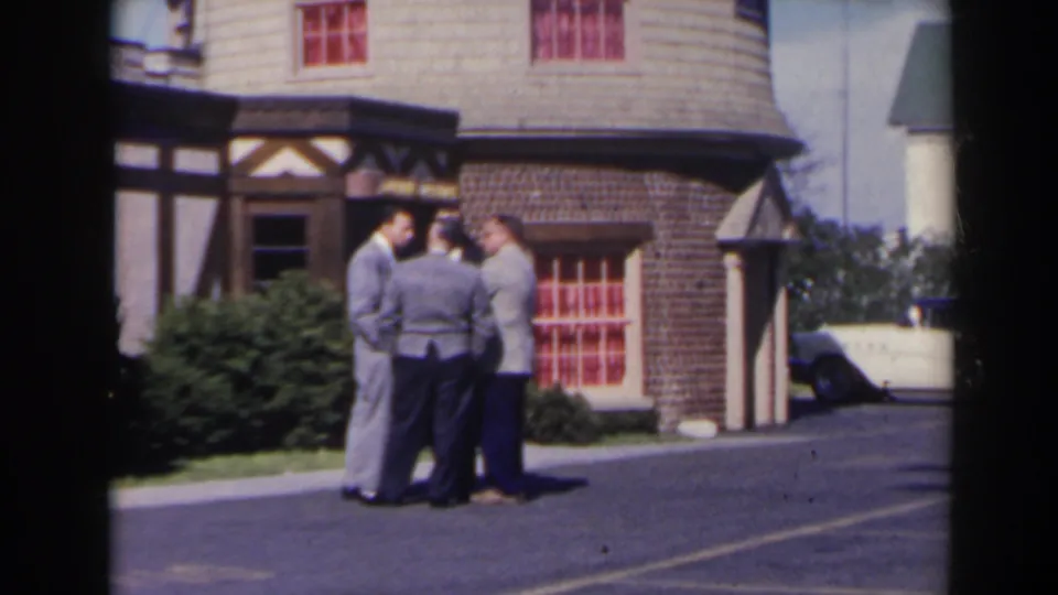 1963 Niagara Falls home-movie frame showing three well-dressed men conversing outside a Tudor-style building with brickwork, red windows, and a parked car nearby.