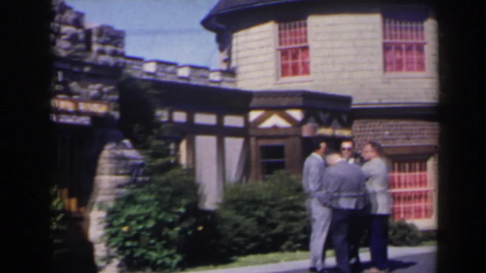 Three men remain grouped outside the castle-like Tudor building, with stonework and red windows visible.