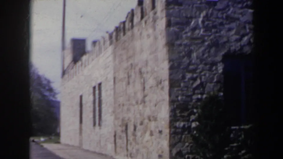 1963 Niagara Falls, Canada frame showing a long stone wall, narrow windows, roadside view, and part of a round tower at right.