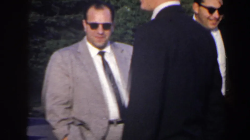 Two adult men in suits and sunglasses stand beside a light-colored car during the 1963 Niagara Falls Canada home movie.