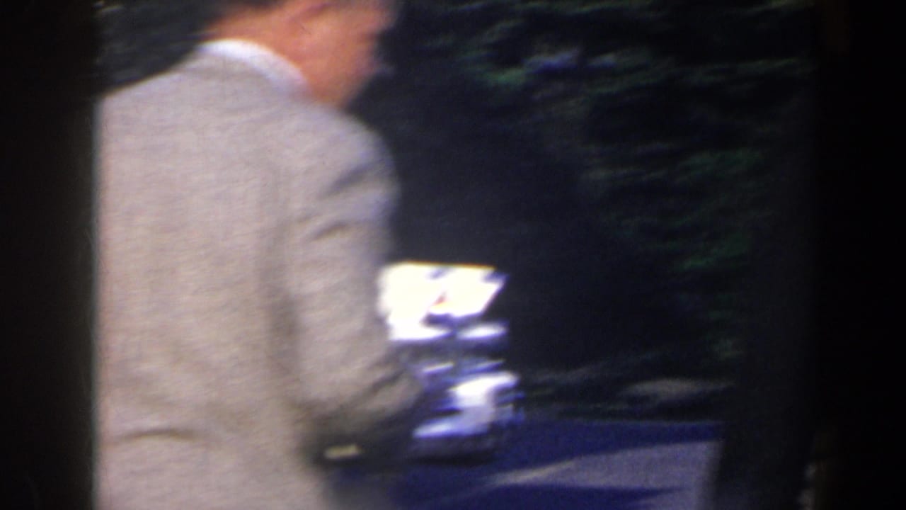 Blurred person in a light jacket walking past trees and a bright outdoor area.