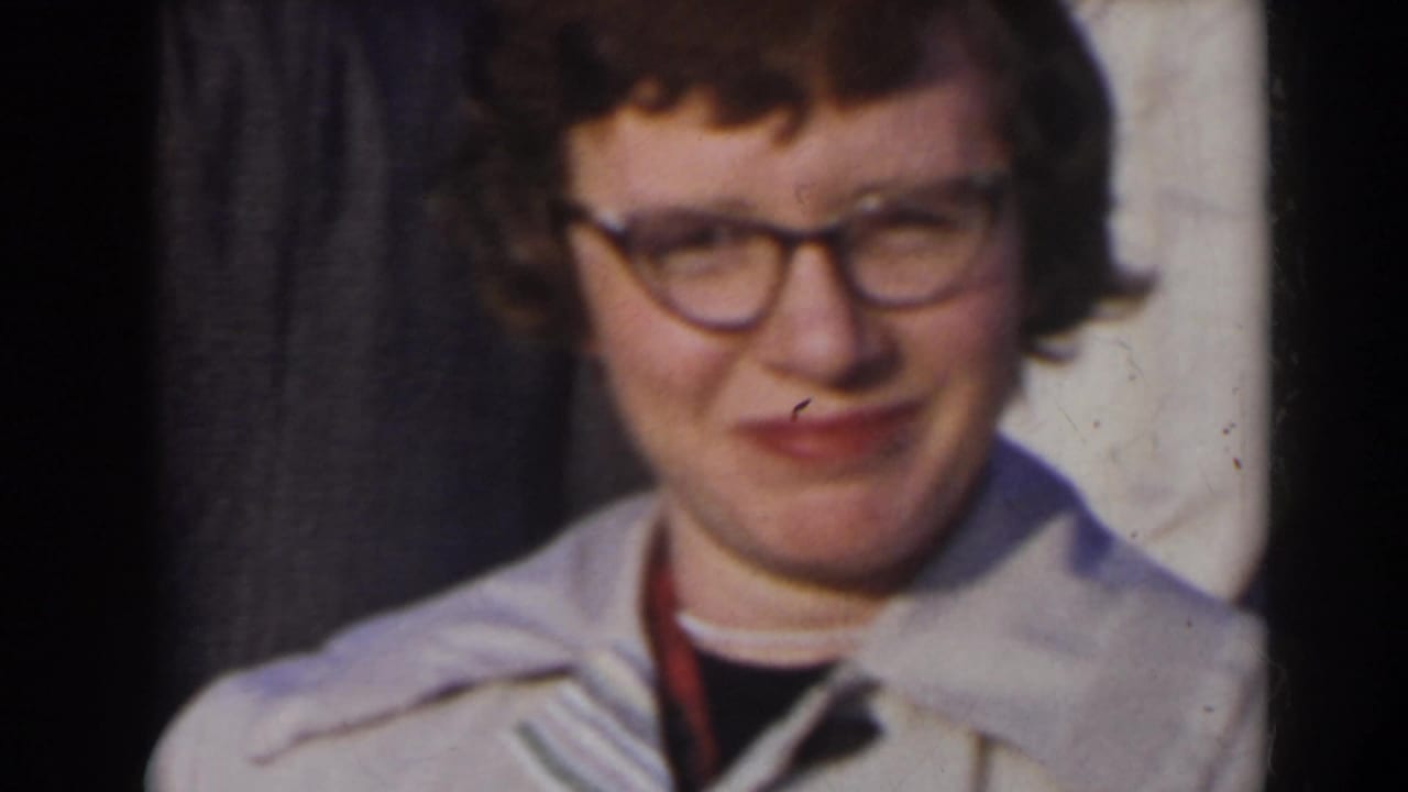 Young woman with glasses and a light-colored jacket standing near trees.