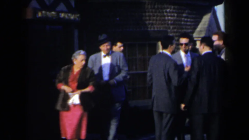 Adults in suits and a woman in a red dress gather outside, with a building and shaded public area visible behind them.