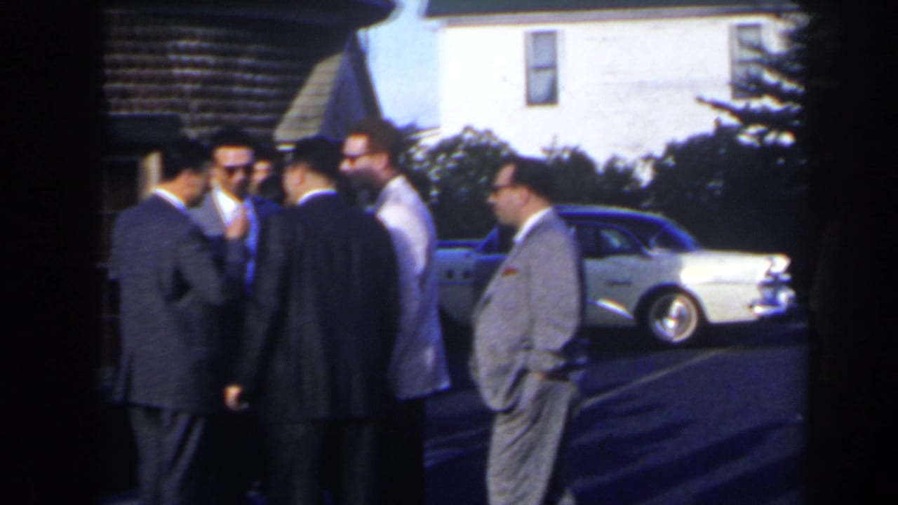 Men in suits converse near parked sedans outside a white building.