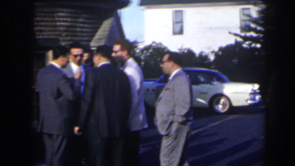 1963 home-movie frame in Niagara Falls, Canada, showing men in suits talking beside mid-century cars in a parking lot.