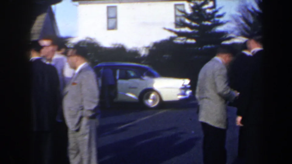 Men in formal jackets gather near a white mid-century sedan in a Niagara Falls, Canada parking lot in 1963 footage.