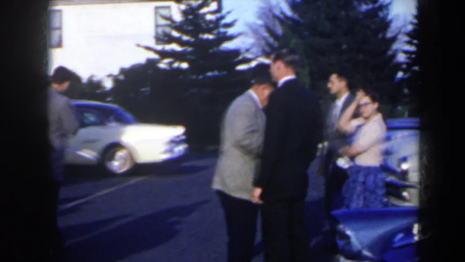 1963 8mm frame shows men in suits and a woman in a light top and patterned skirt talking near cars in Niagara Falls, Canada.