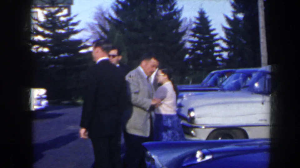 A man in a light suit and a woman in a patterned skirt appear to talk beside a blue car in 1963 Niagara Falls home-movie footage.