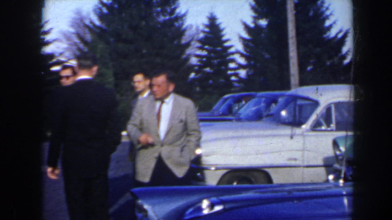 Adults stand near parked mid-century cars outside a building.