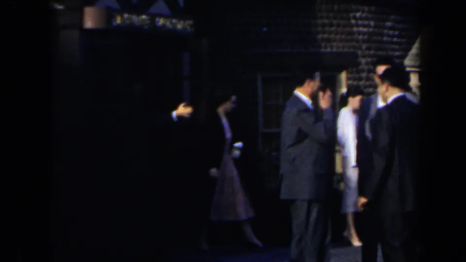 A shaded 1963 Niagara Falls frame shows suited men and women in light coats talking outside a building.