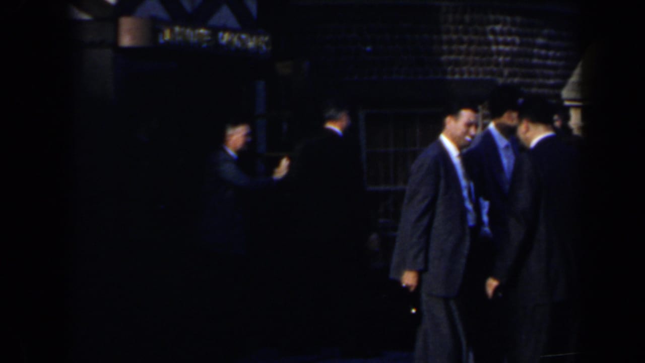 1963 8mm home-movie frame of suited men standing outside at night in Niagara Falls, Canada, near a dim building and partial sign.