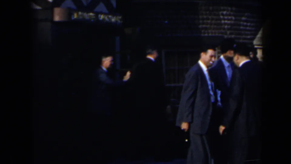 1963 8mm home-movie frame of suited men standing outside at night in Niagara Falls, Canada, near a dim building and partial sign.