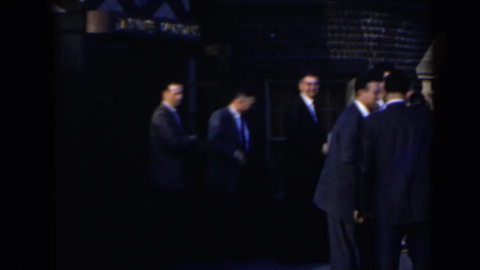 Adults in suits appear to converse outside at night in 1963 Niagara Falls home-movie footage, with a dim facade behind them.