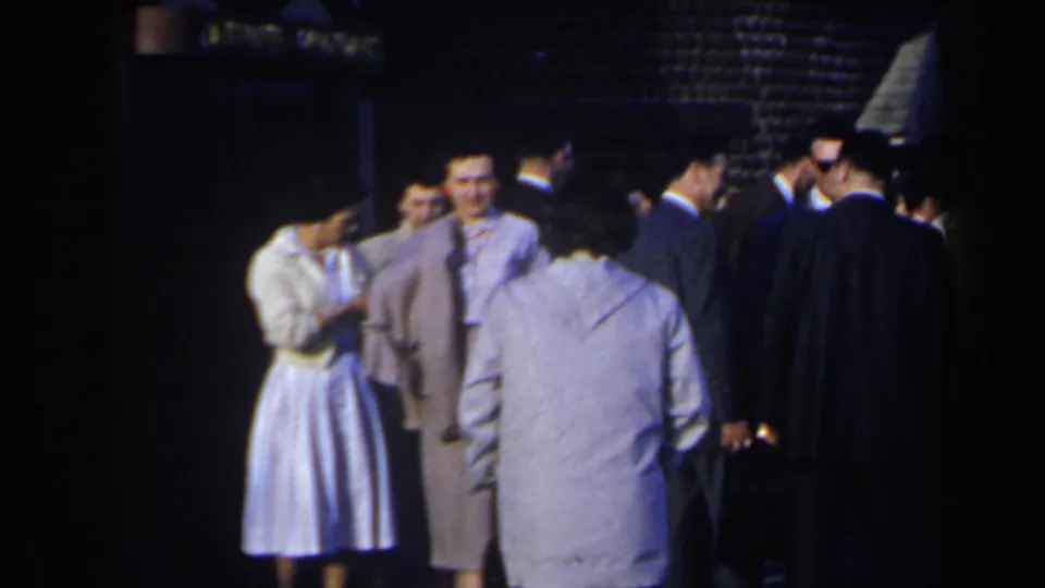 A woman in a pale dress stands near suited men in a nighttime 1963 Niagara Falls home-movie scene, with faces partly visible.