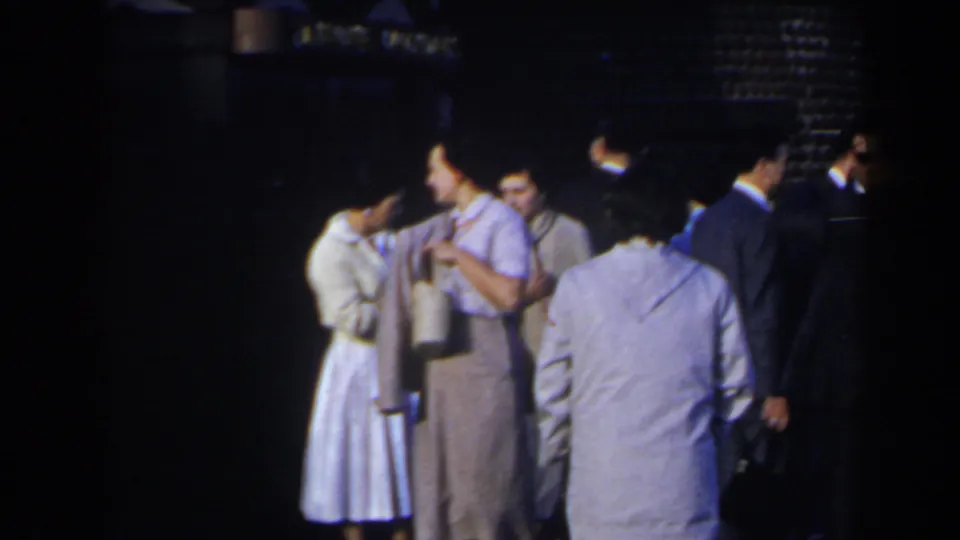1963 Niagara Falls home-movie frame showing adults gathered outside, including a woman in a light dress and men in suits near an “AIR CONDITIONED” sign.