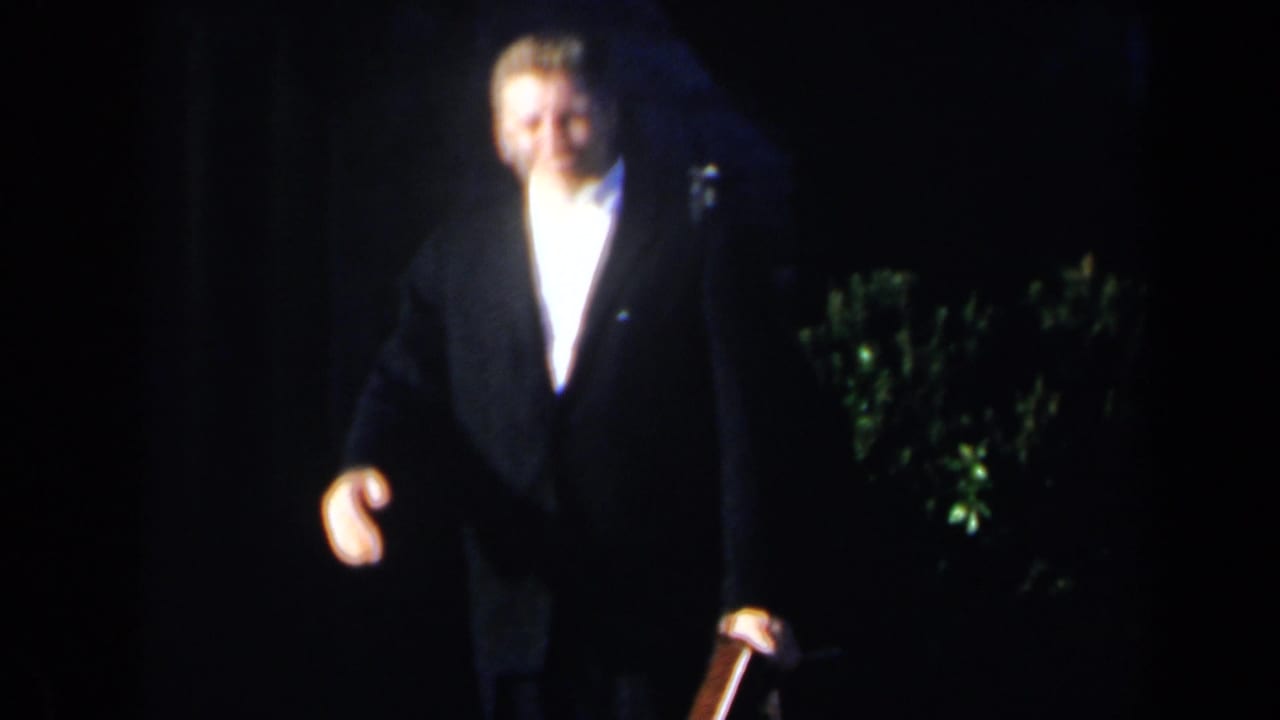 A man in a dark suit walks forward in a dimly lit outdoor setting.
