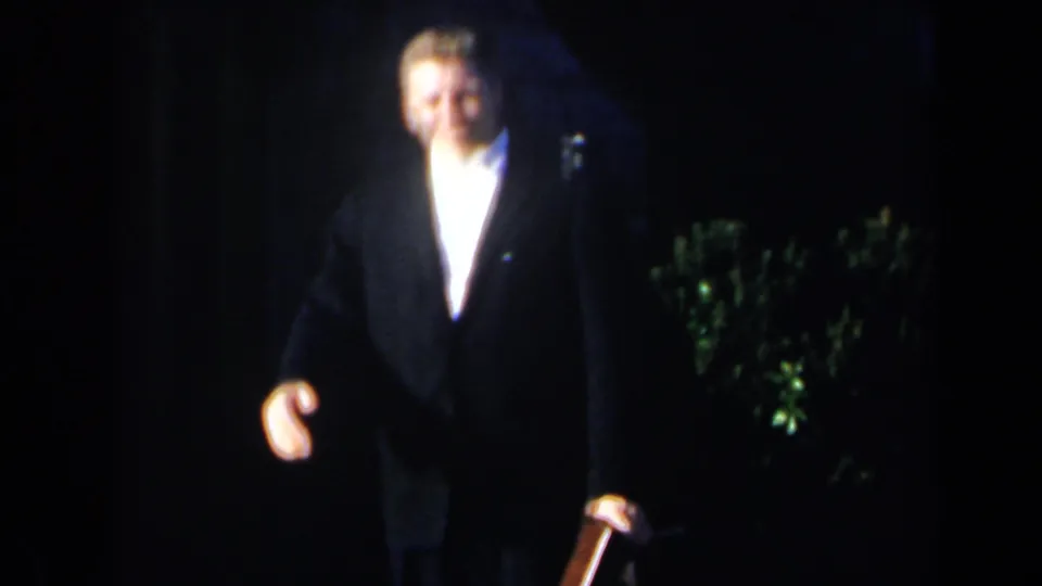 1963 Niagara Falls frame of a smiling man in a dark suit and white shirt walking forward, with greenery and dark surroundings behind him.