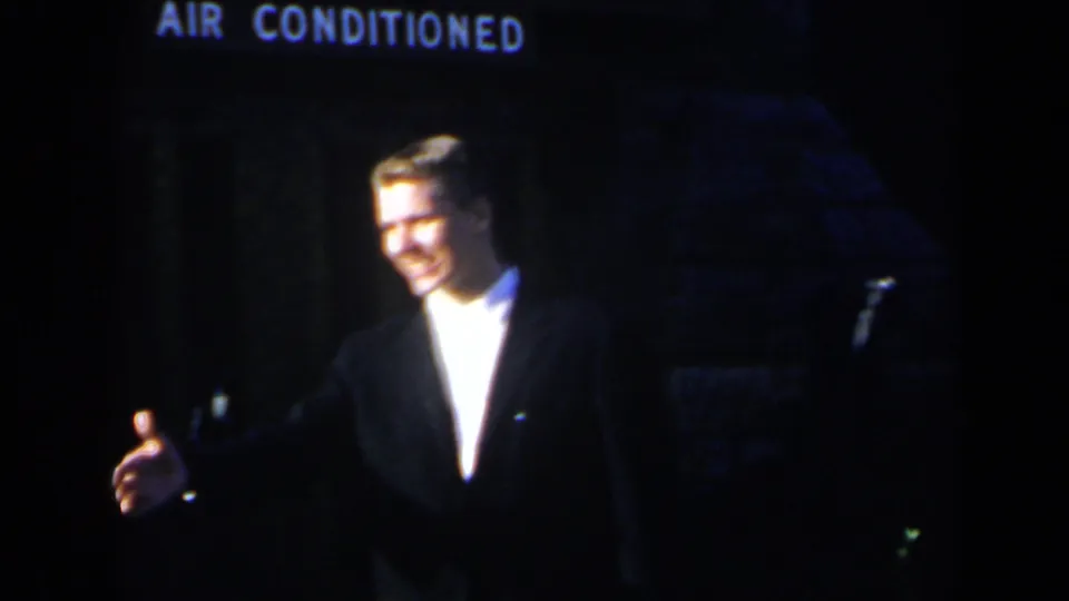 1963 Niagara Falls home-movie frame showing a smiling suited man extending his hand or gesturing in a dim outdoor scene.