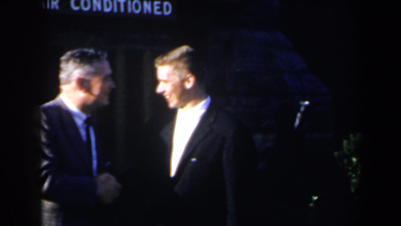 Two suited men shake hands on a dim stage beneath a partial air-conditioned sign.