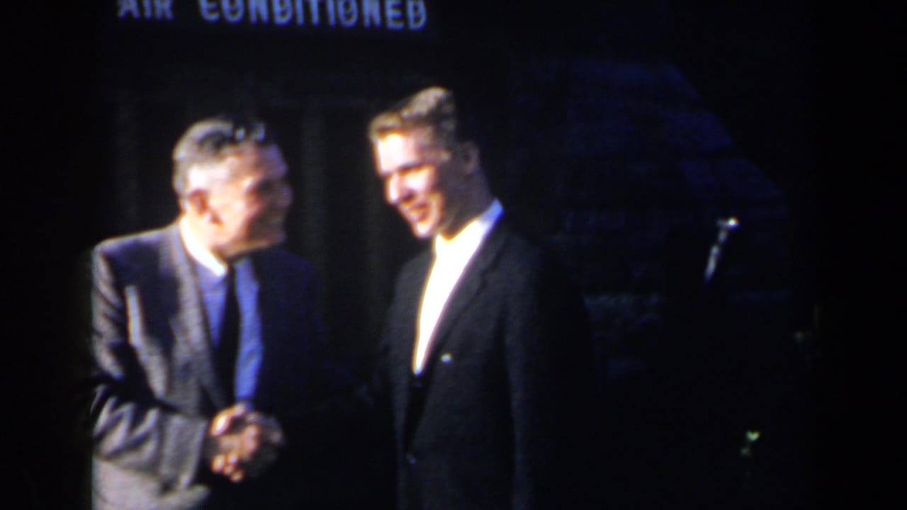 The suited men face each other and smile during a formal handshake, with dim stage lighting and signage behind them.