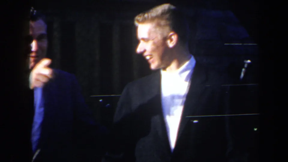 A young suited man smiles on the dim stage as another man appears partly in shadow, with a microphone stand visible nearby.
