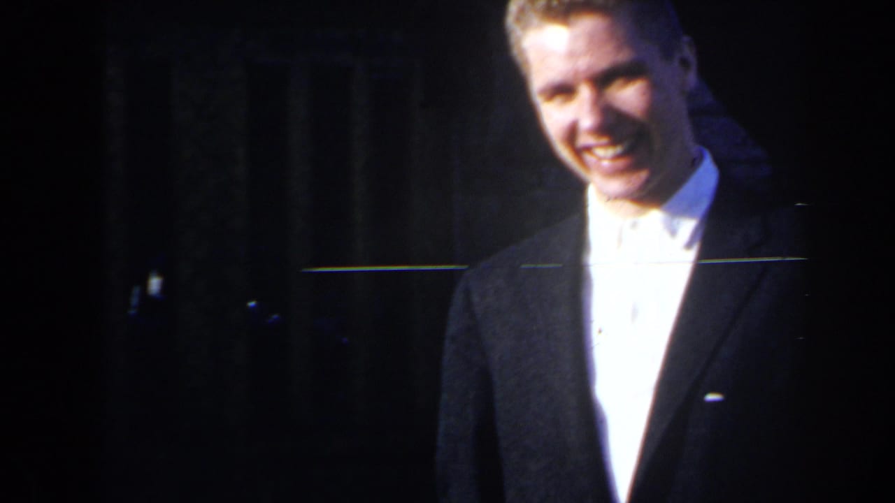 Young man in a dark suit smiles toward the camera in a dim setting.