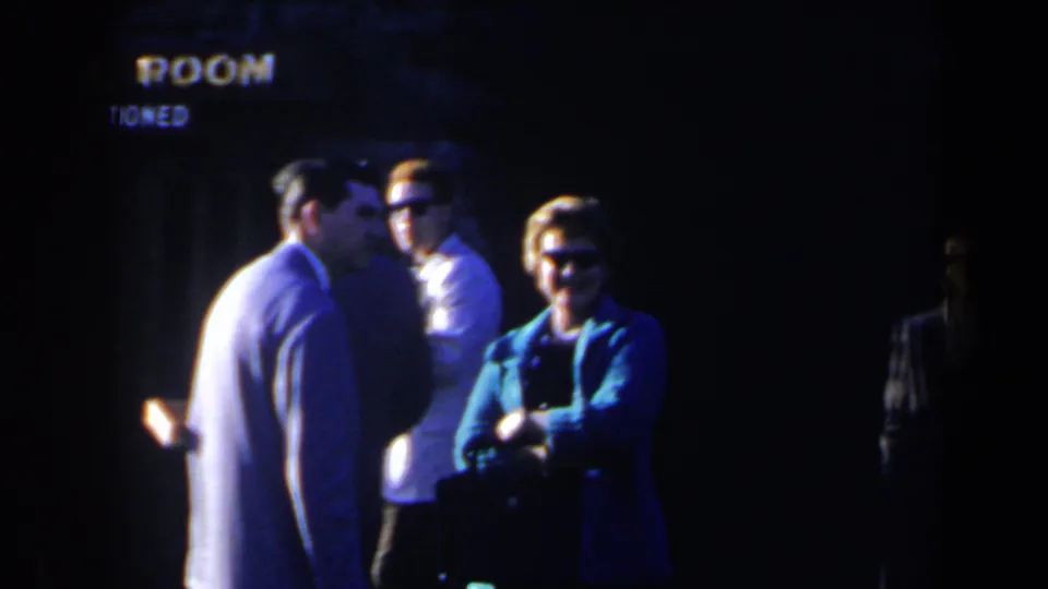 Adults in suits and sunglasses stand near partial room signage in the Niagara Falls 1963 home-movie scene.