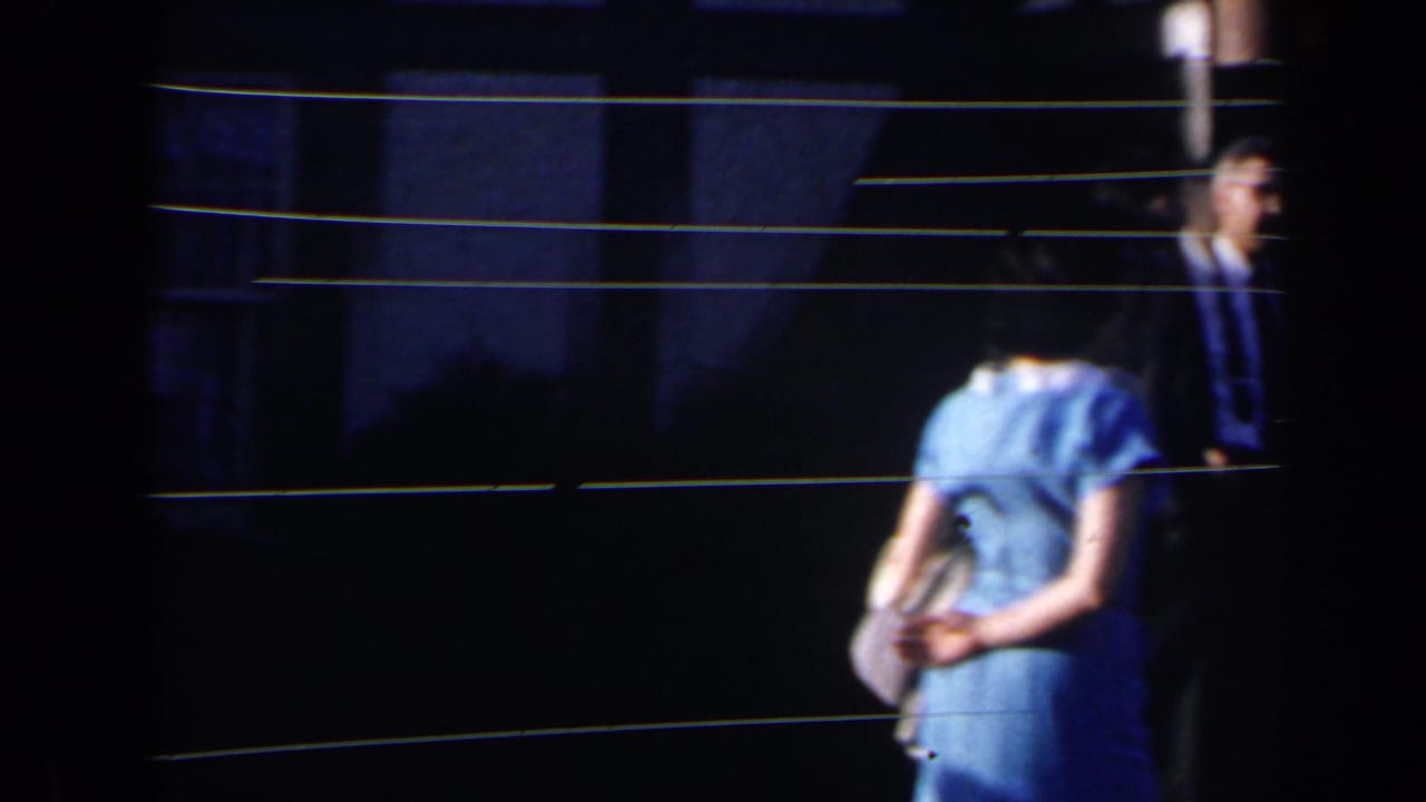 Woman in a light blue dress stands near a suited man by a dark entrance.