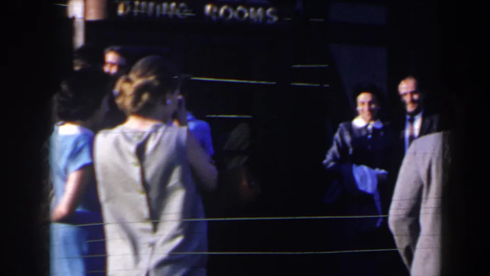 1963 Niagara Falls home-movie frame showing adults in dresses and suits gathered near a dark dining-room entrance.