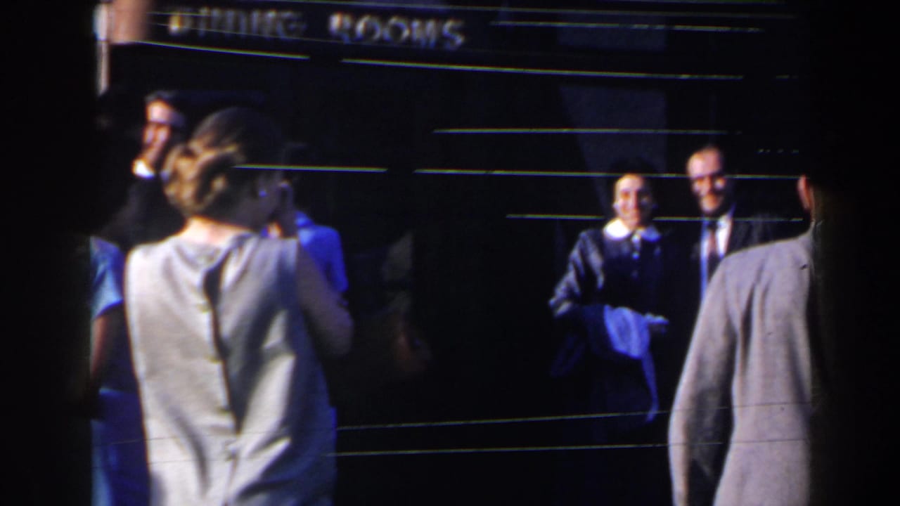 1963 Niagara Falls home-movie frame showing adults in suits and dresses near a dining rooms entrance.